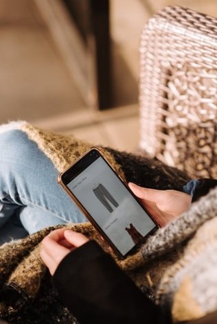 A woman wearing a black shirt and blue jeans is sitting in a straw chair as she scrolls through Amazon to look at grey pants and a red top while she snuggles with her woolen scarf