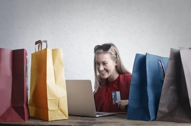A blonde woman in a red shirt with black and brown sunglasses on her head is smiling at her laptop screen with her credit card in her hand, presumably ordering items online as she is surrounded by red, yellow, blue, and grey gift bags from her previous online purchases